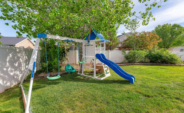 Wooden swing set with green saucer swing and blue slide installed in a fenced backyard