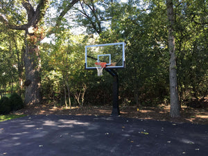 In-ground basketball hoop with a tempered glass backboard on a paved backyard court surrounded by trees
