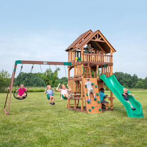 Children swinging, climbing the rock wall, and sliding on a cedar Backyard Discovery swing set with a green wave slide in a grassy yard