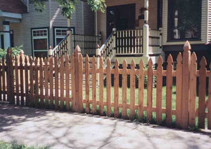 Cedar gothic picket fence along a sidewalk in front of a Victorian home with a wood porch
