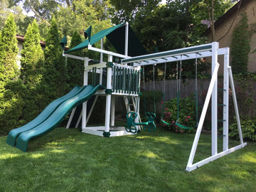 White vinyl playset with green slide, swings, and green canopy in a fenced backyard