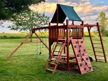 Wooden swing set with green canopy, climbing wall, slide, and swings in a backyard at sunset